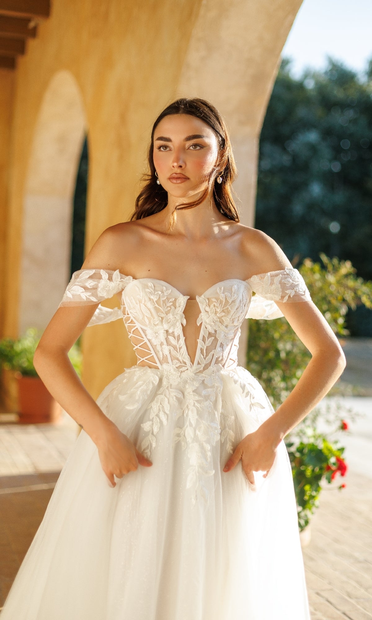 A woman stands outdoors near an arched walkway wearing the Tina Holly TW480, a long formal off-the-shoulder white bridal gown with a floral lace corset bodice.